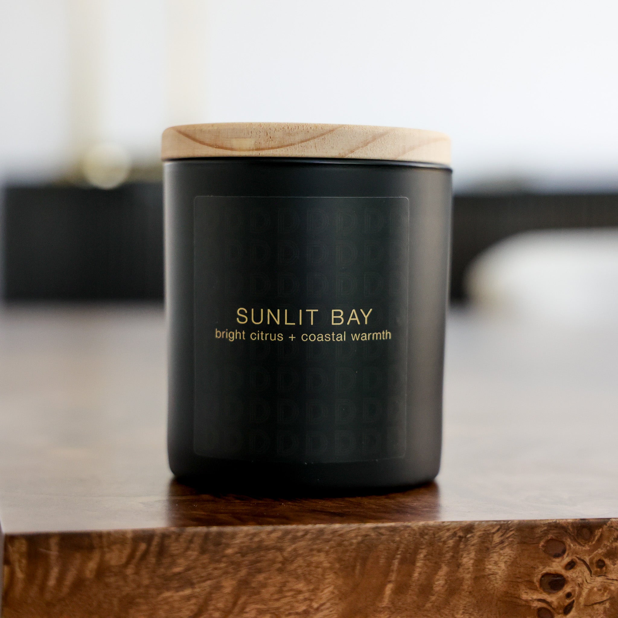 A black candle jar with a light wood lid, labeled 'SUNLIT BAY bright citrus + coastal warmth', sitting on a wooden surface with a blurred indoor background.