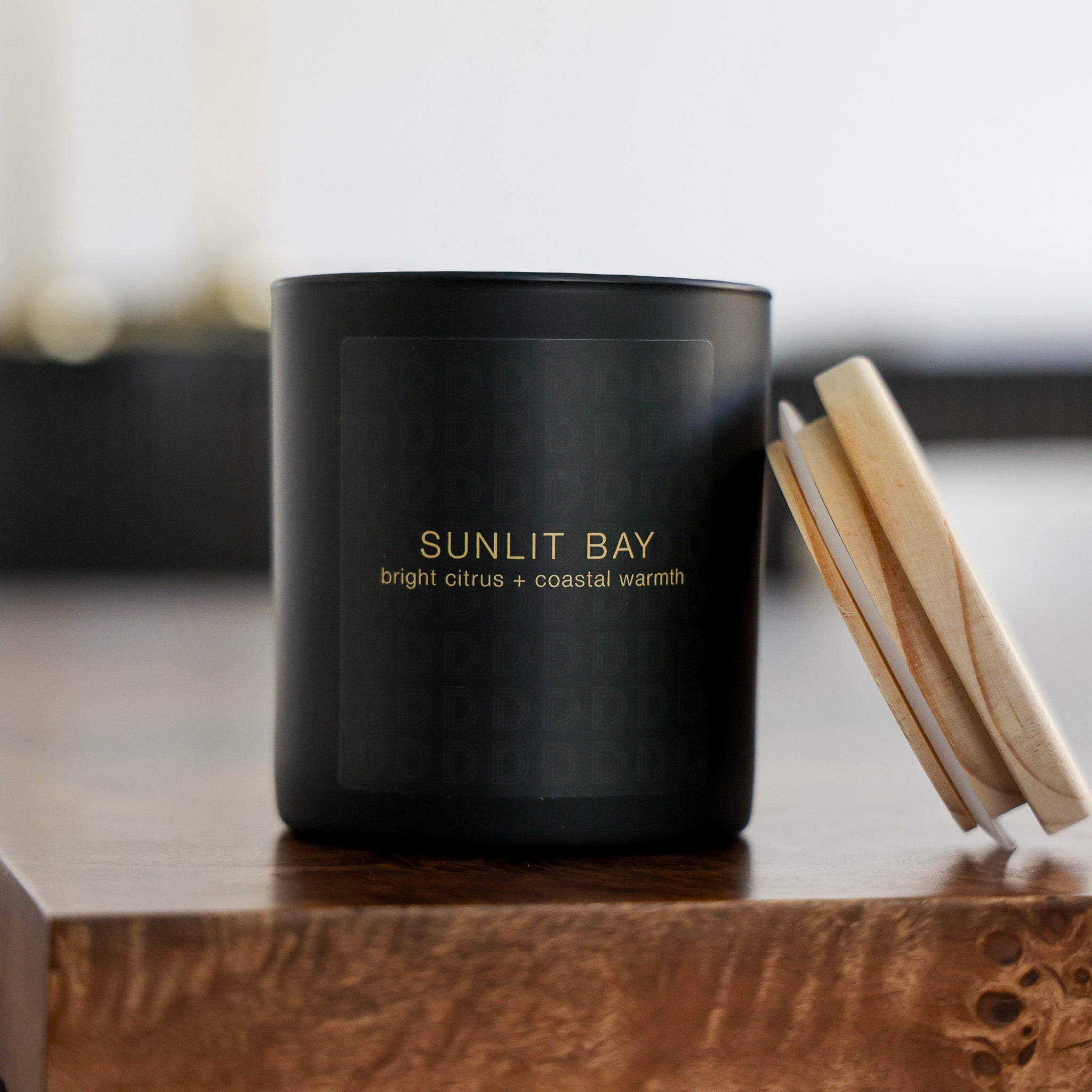A matte black candle jar labeled 'SUNLIT BAY' sits on a wooden surface with its light wood lid leaning against it. The background is softly blurred, creating a warm, cozy indoor atmosphere.