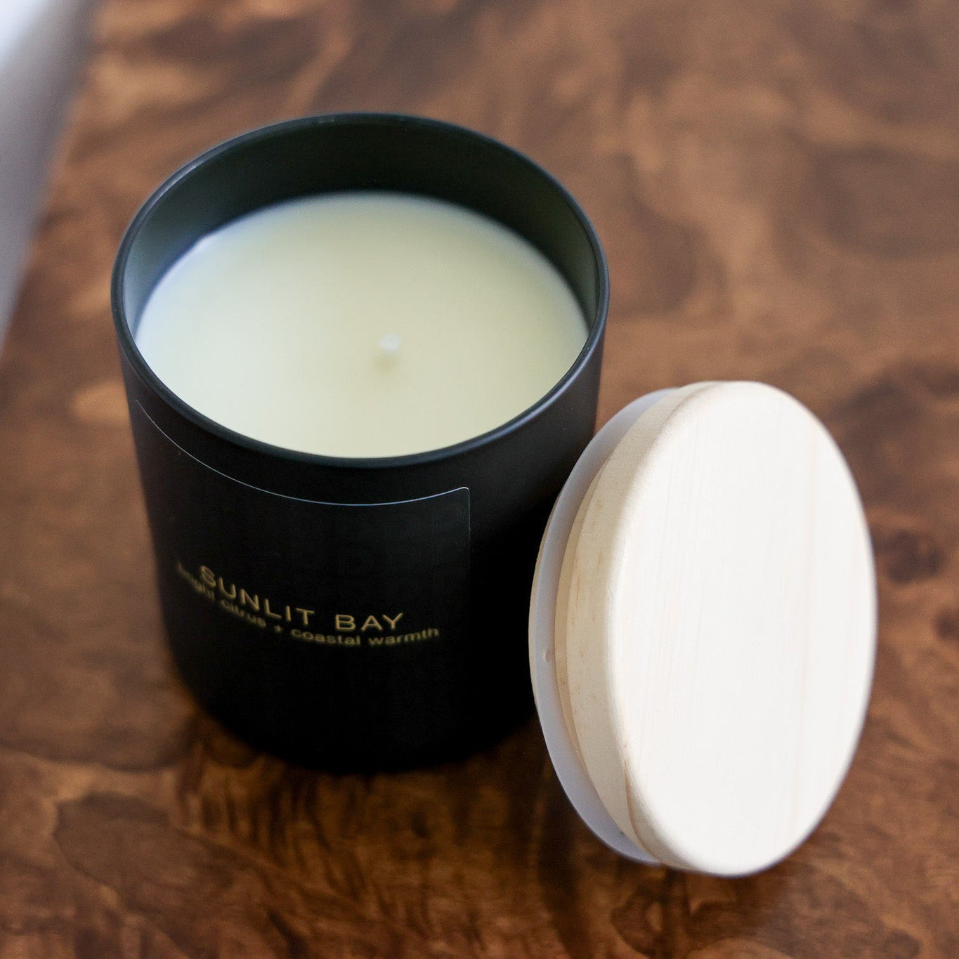 An open black candle jar with pale yellow wax sits on a wooden surface next to its white lid. The jar is labeled 'BUNLIT BAY'.