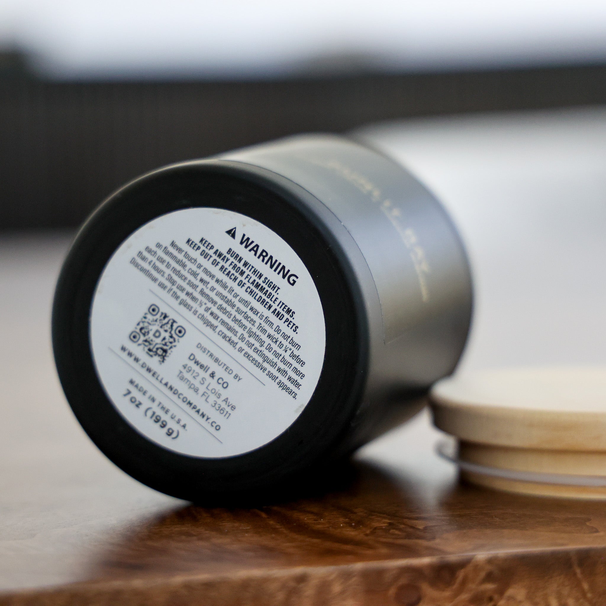 A close-up of a black insulated tumbler lying on its side on a wooden surface, showing the printed warning label on the bottom. A stack of light wood lids is visible in the background.