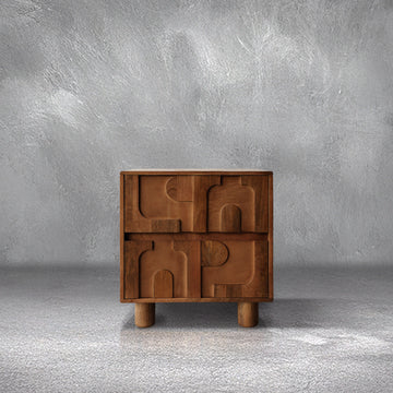 Funky Nightstand in warm mango wood with 3D carved drawer fronts and cylindrical feet, beside a bed