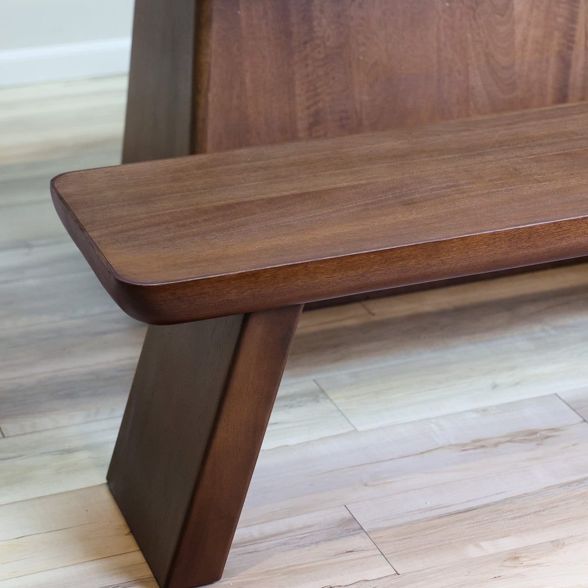 A close-up of a modern wooden bench with a rich dark brown finish, featuring a thick slatted backrest and angled legs, set on a light wood plank floor.