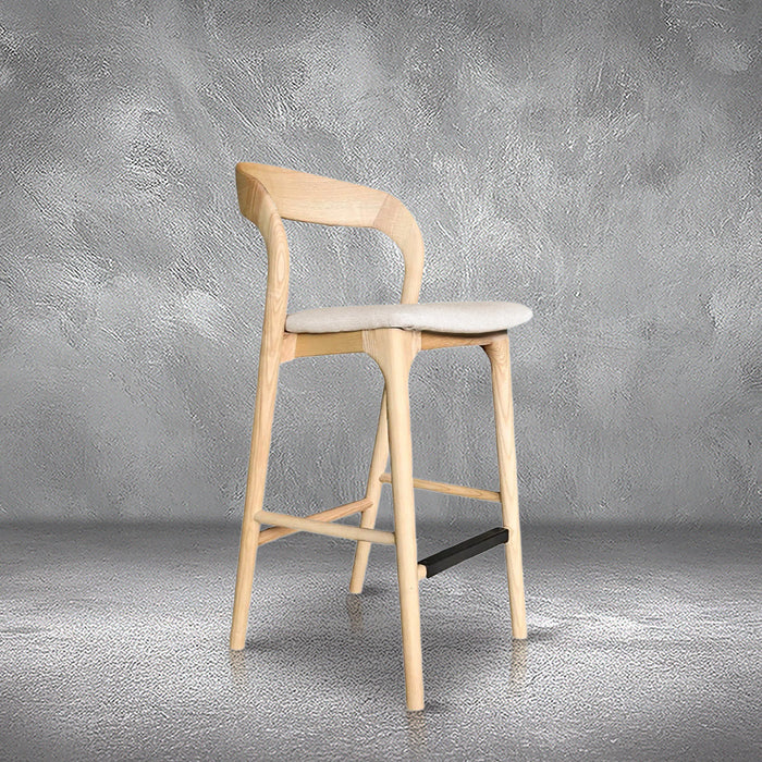 Joan counter stool in light ash wood with a curved wraparound back, upholstered beige seat, and footrest, kitchen counter
